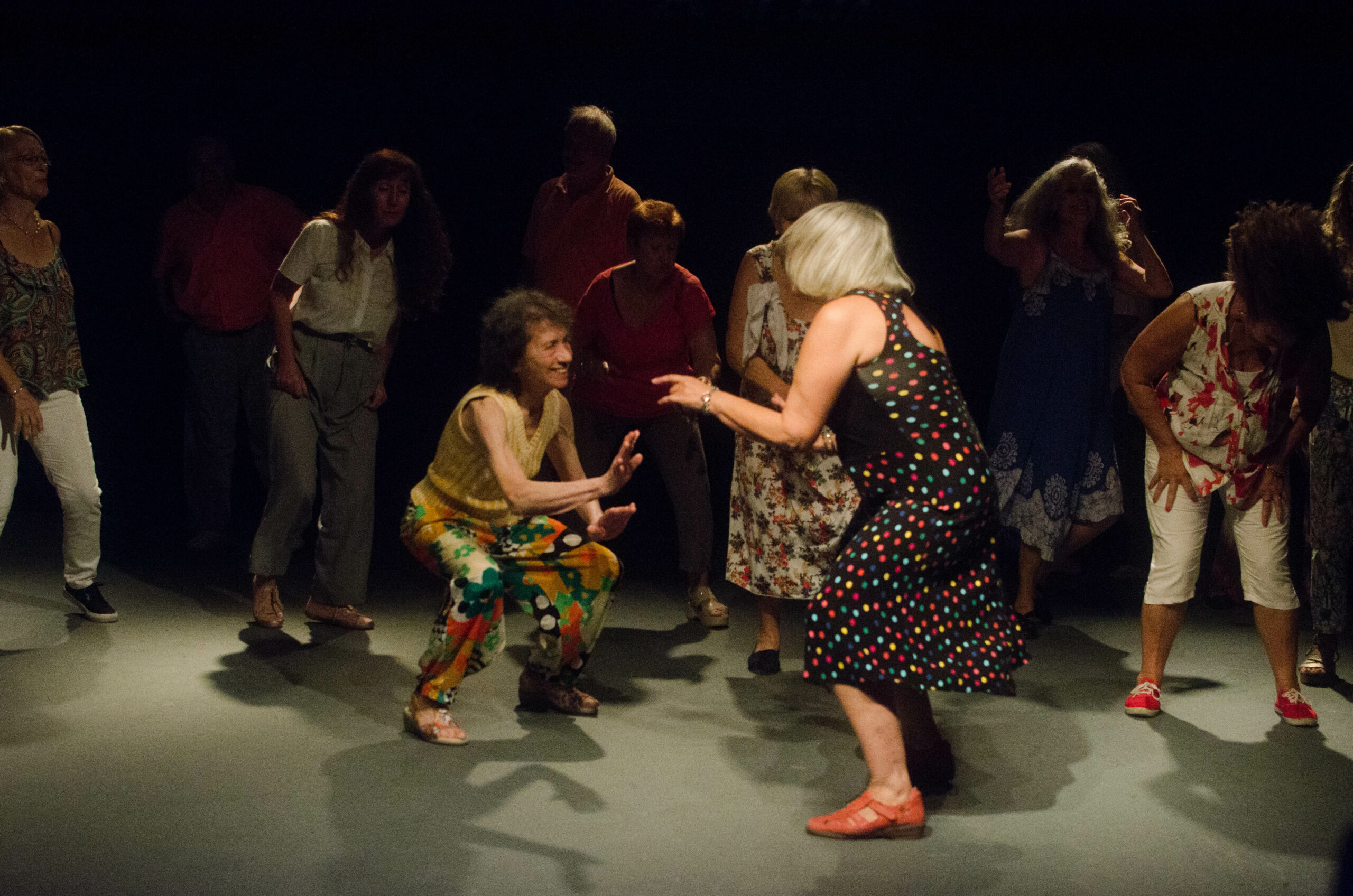 Grupo de adultos mayores en un escenario donde se ven dos mujeres protagonizando la escena con una danza entre ellas mientras el resto las rodea.